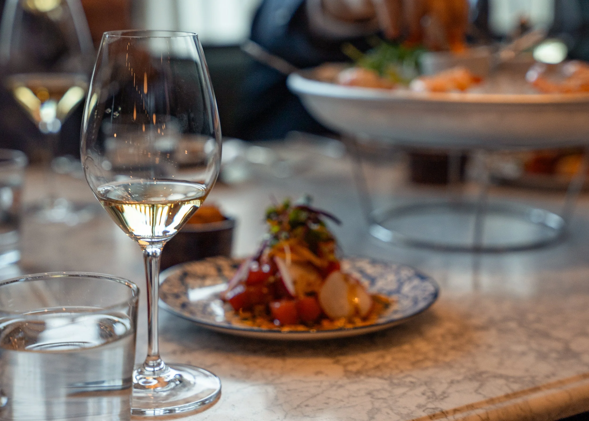 A glass of white wine, a glass of water, and a plate of food on a marble table at a restaurant or event.