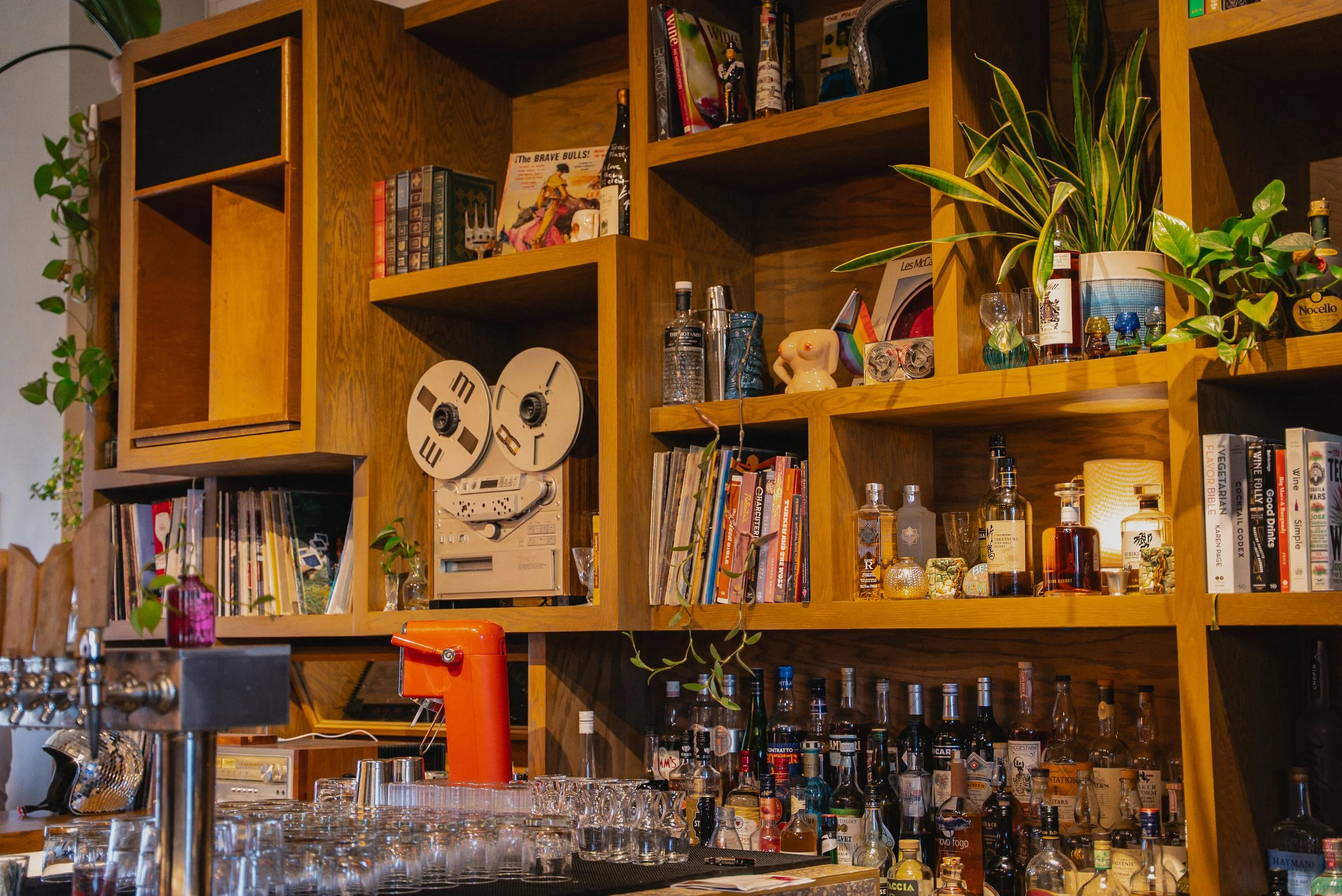 View of a wooden bar with shelves filled with books, bottles, plants, and decorative items. A vintage reel-to-reel tape recorder is on the shelves, along with vinyl records and various liquor bottles. Glassware is arranged on the bar counter.