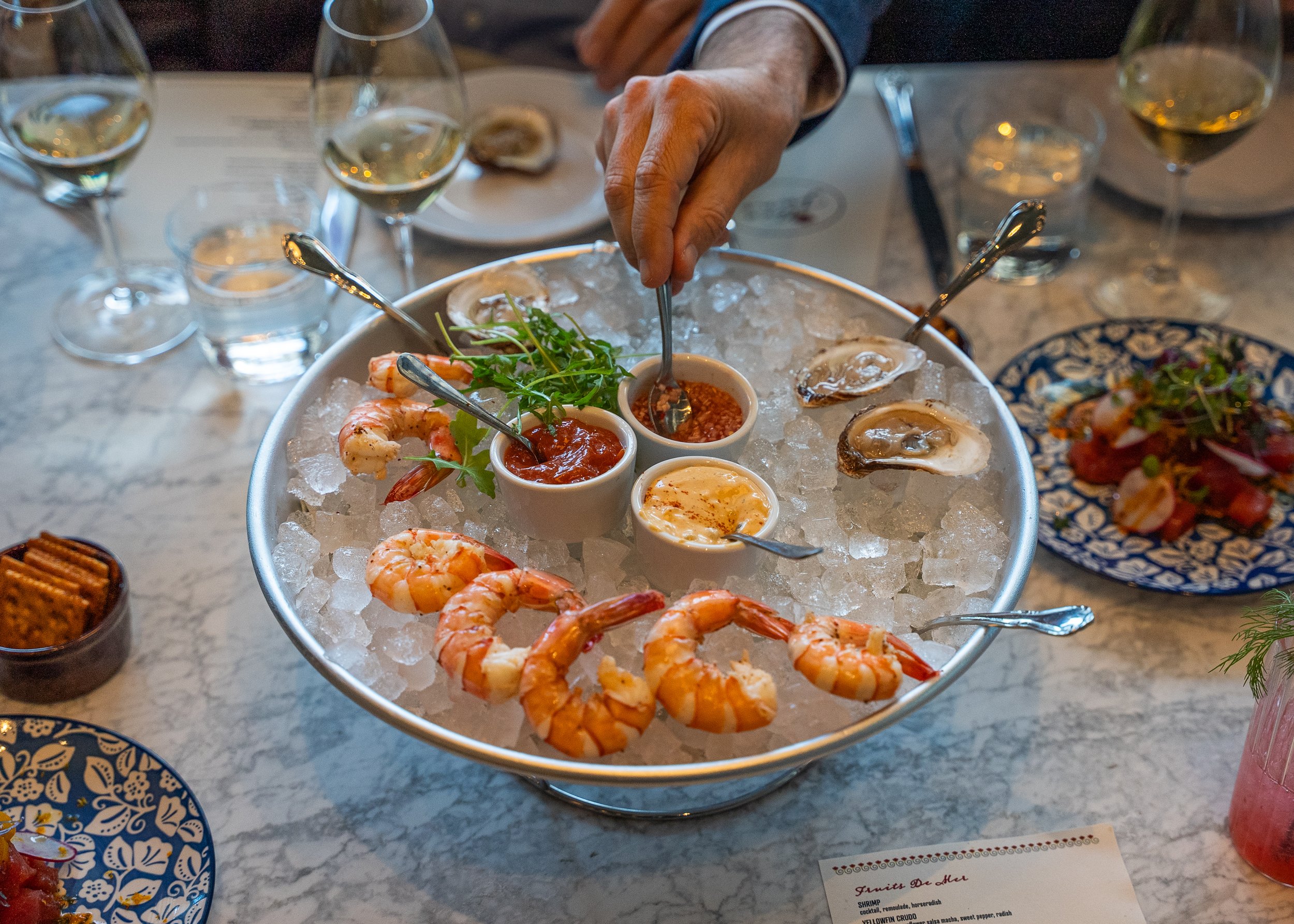 A platter of shrimp, oysters, and dipping sauces on ice at a restaurant. There are several glasses of white wine and a side dish on the table.