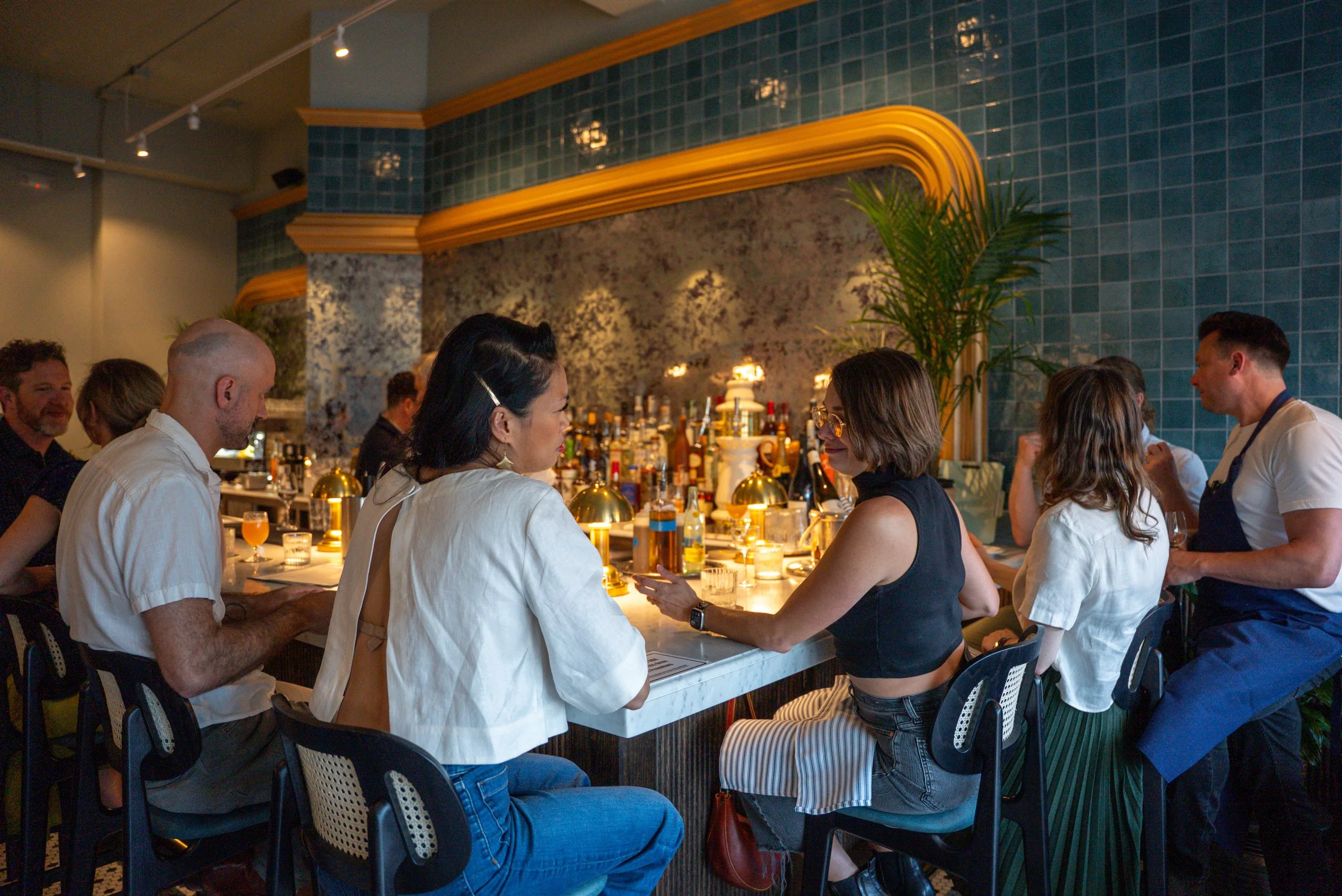 A group of people sitting at a bar in a restaurant, engaging in conversation. The bar is decorated with bottles, glasses, and warm lighting, with a marble wall and green tiles in the background.