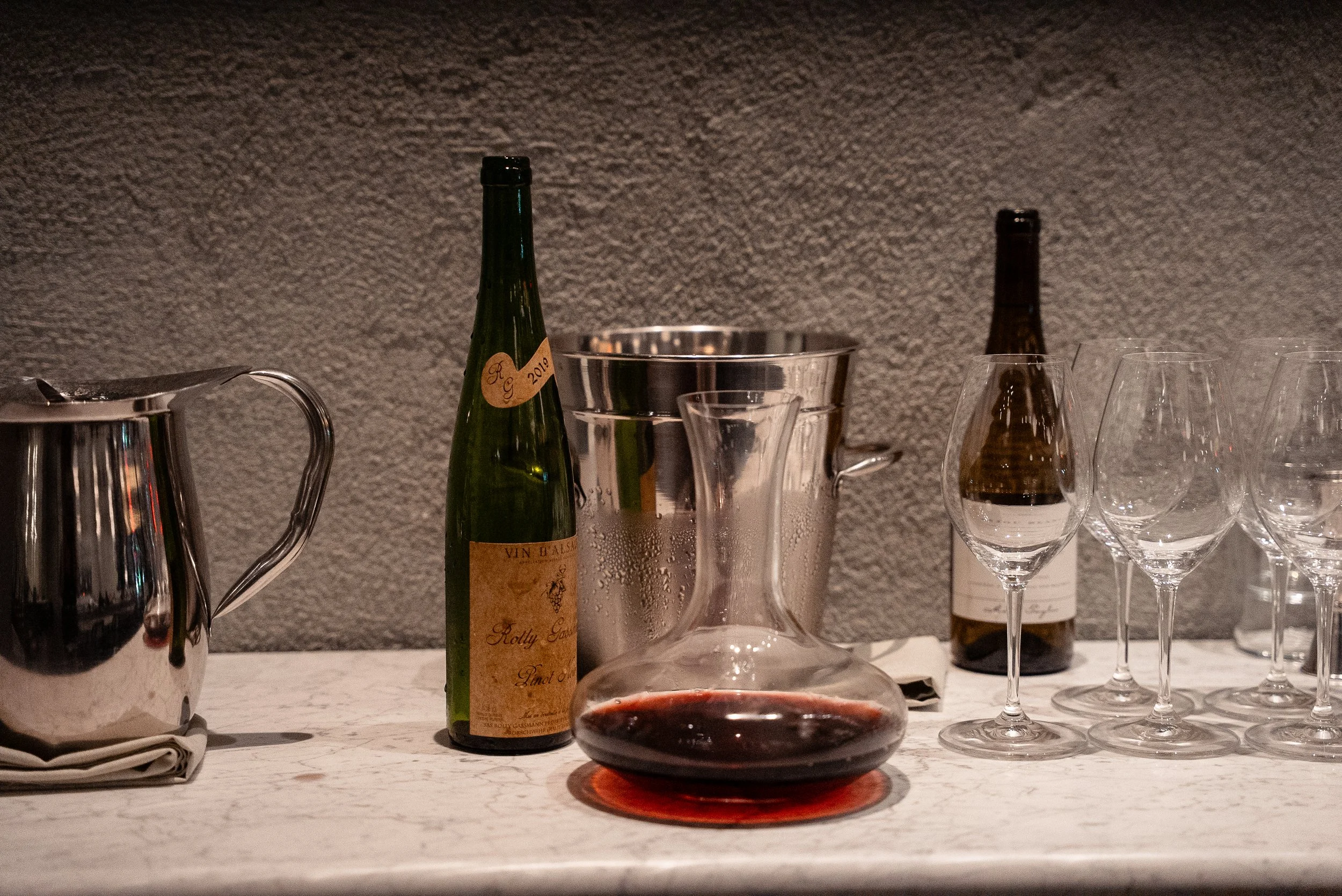 Table setup with a partly filled wine decanter, wine bottle, wine glasses, a metal ice bucket, a glass pitcher with condensation, and a wine bottle in the background against a textured gray wall.