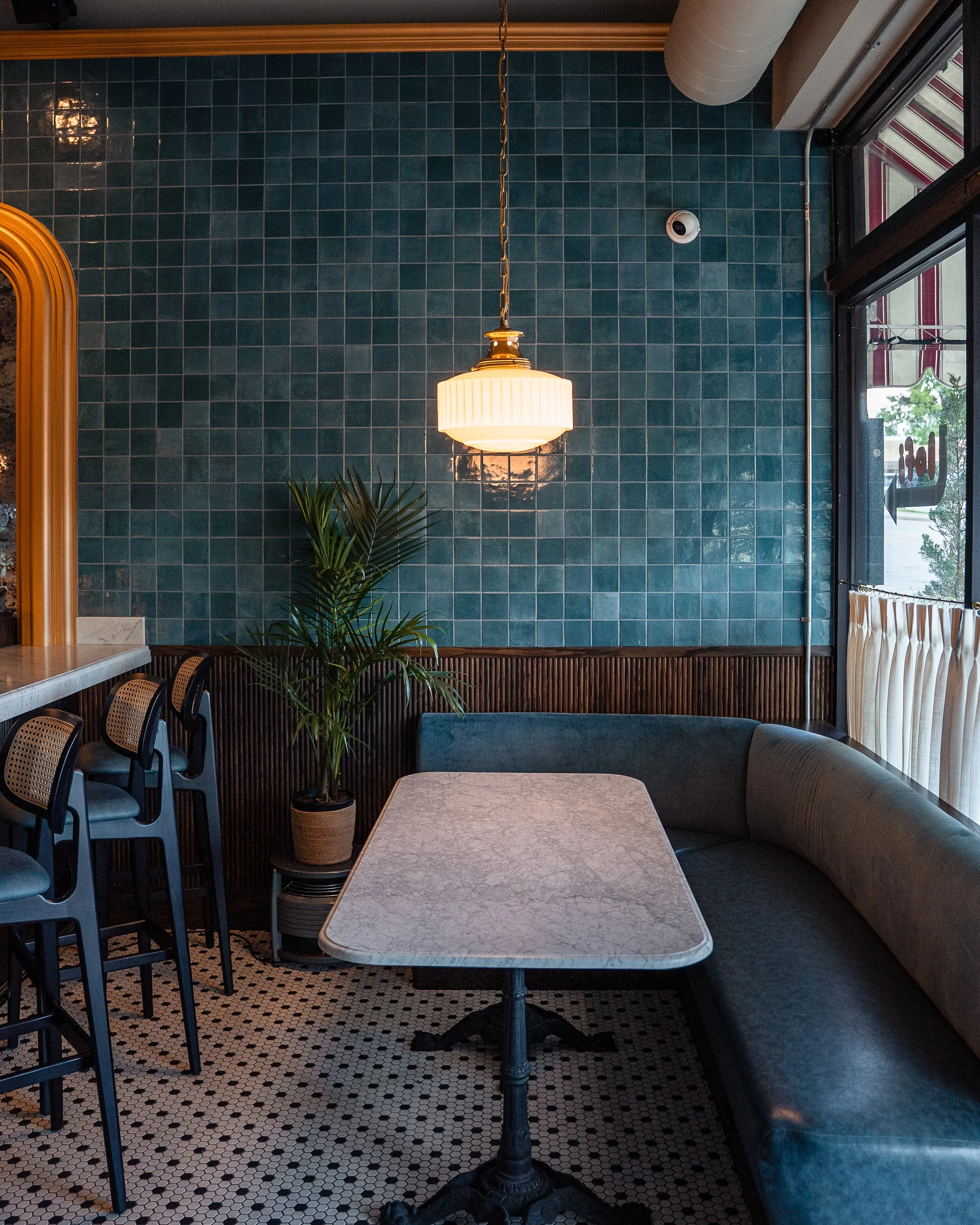 A cozy cafe corner with a marble table, a curved blue velvet booth, a potted plant, teal tiled wall, hanging pendant light, and a window with curtains.