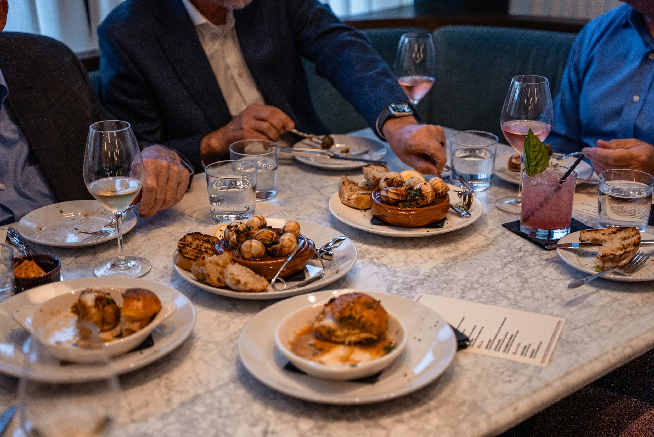 People dining at a restaurant with plates of food, bread, and drinks on a marble table.