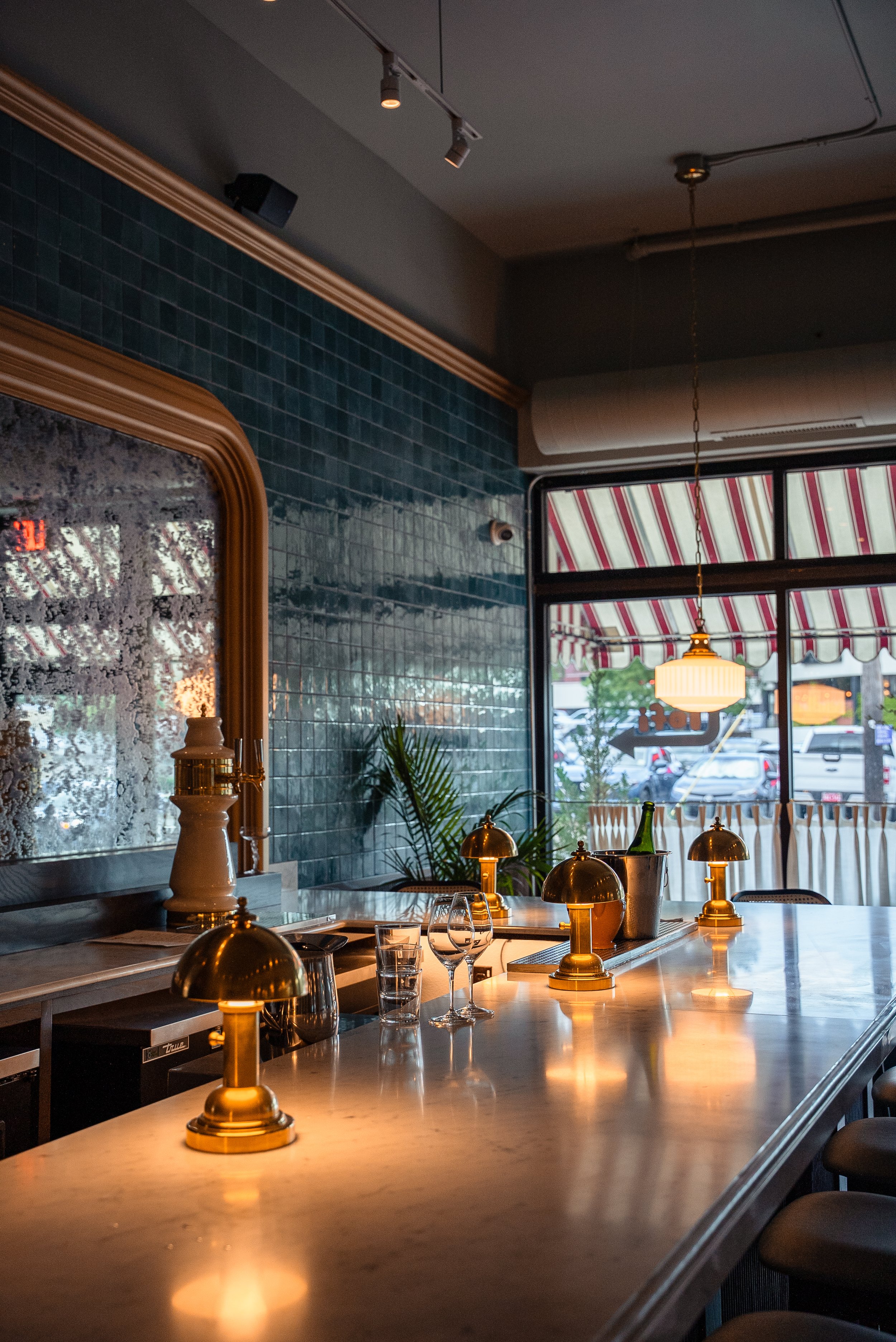 Interior of a stylish bar or restaurant with a marble countertop, small gold desk lamps, glasses, and a bottle of champagne. Large windows with striped red and white awnings outside.