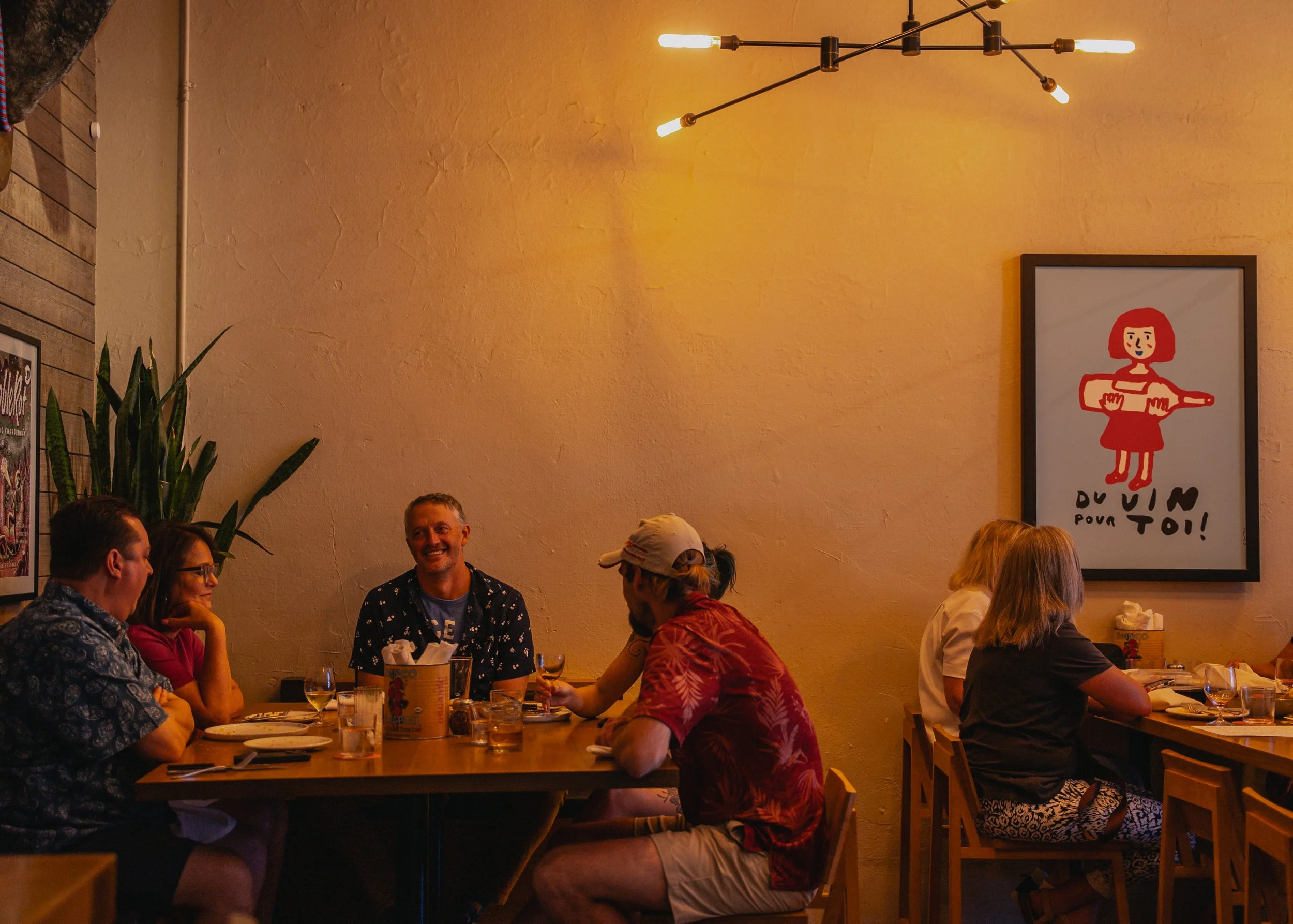Group of people enjoying dinner in a cozy restaurant with warm lighting, a large plant, and colorful artwork on the walls.