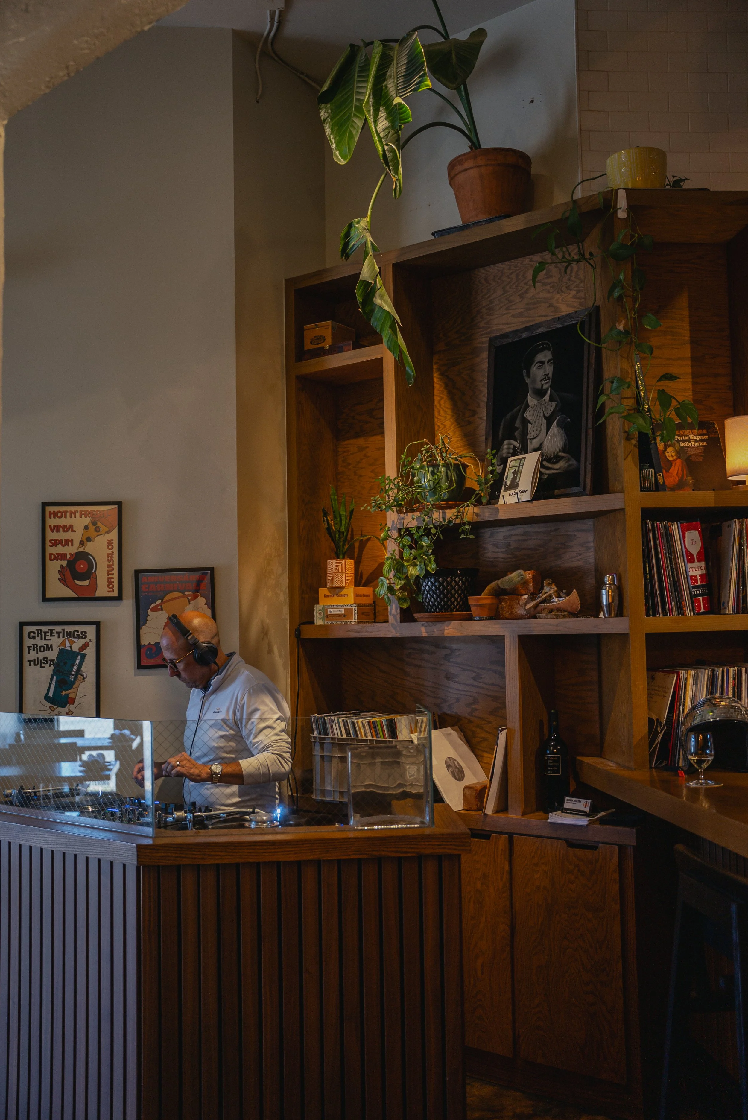 A DJ wearing headphones and glasses, dressed in white, is operating turntables inside a room with wooden shelving filled with books, records, plants, and decorative items.