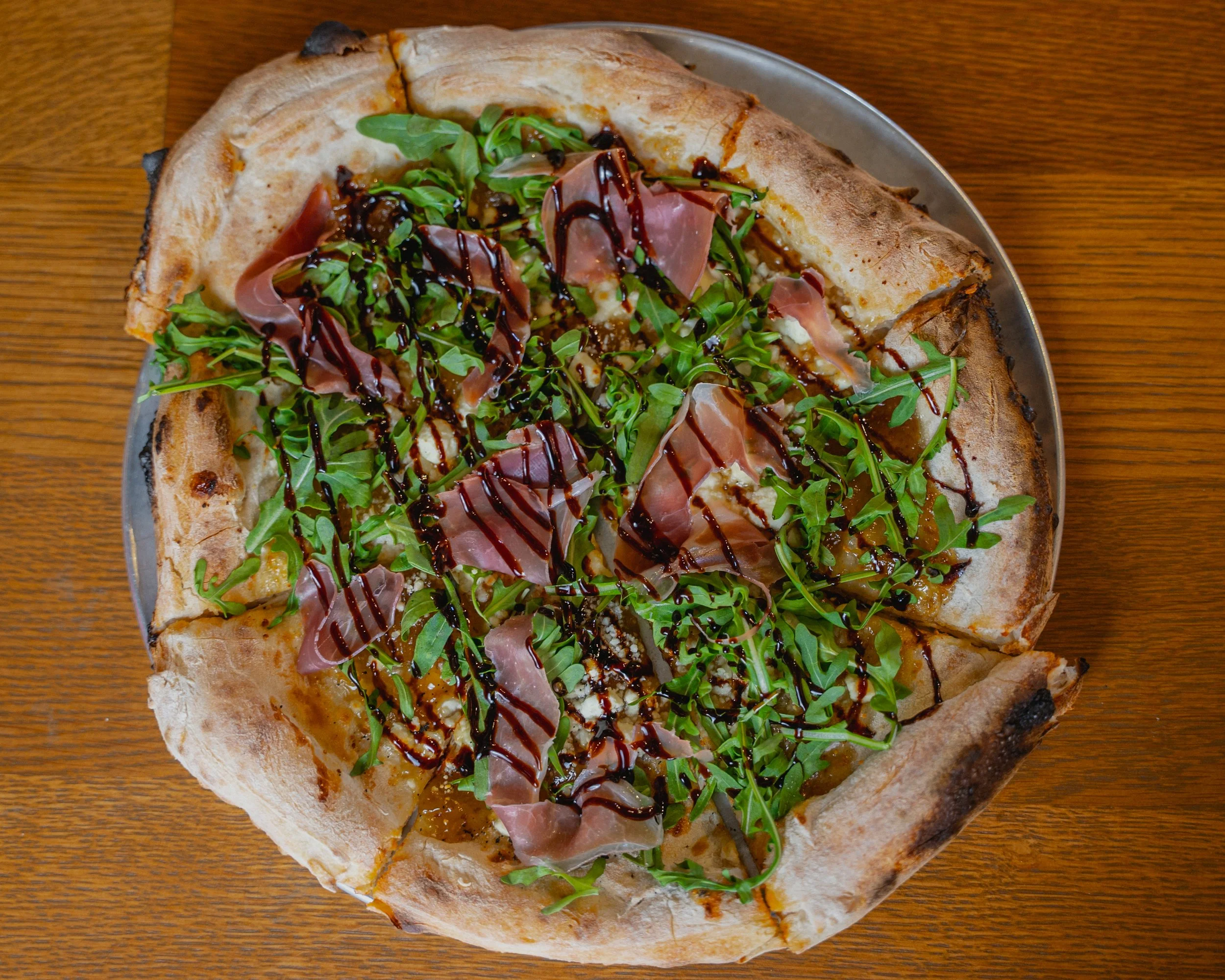 A pizza with a crust, topped with arugula, prosciutto, balsamic glaze, and cheese, on a silver tray on a wooden table.