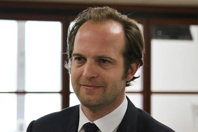 A man with light skin, brown hair, and a slight smile, wearing a black suit, white shirt, and dark tie, standing indoors with large windows in the background.