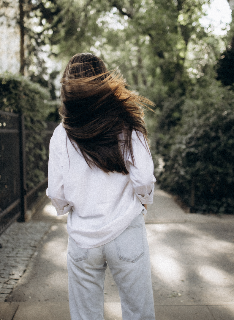 A woman with long, brown hair facing away from the camera, standing on a path in a green park during the daytime.