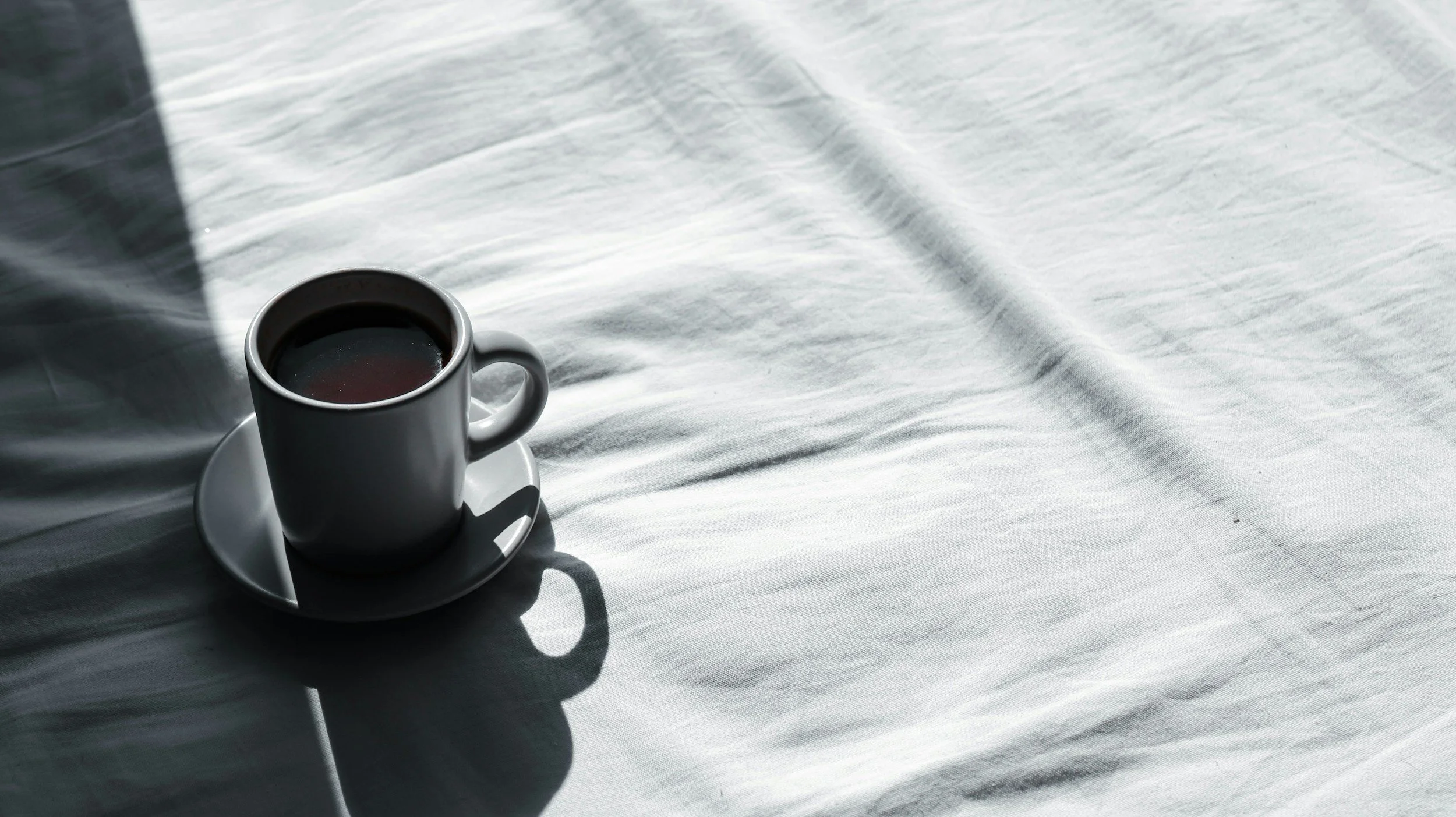 A white coffee cup filled with dark coffee sitting on a matching white saucer on a white fabric surface, with shadows cast on the fabric.