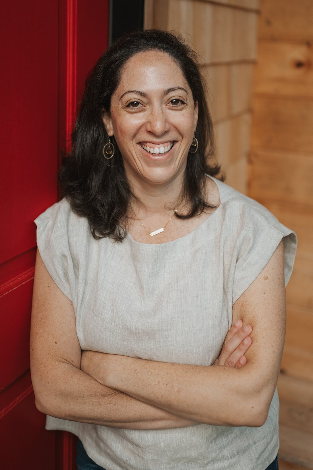 A smiling woman with dark hair, wearing a beige sleeveless top, gold earrings, and a gold necklace, standing with her arms crossed next to a red wall and wooden background.