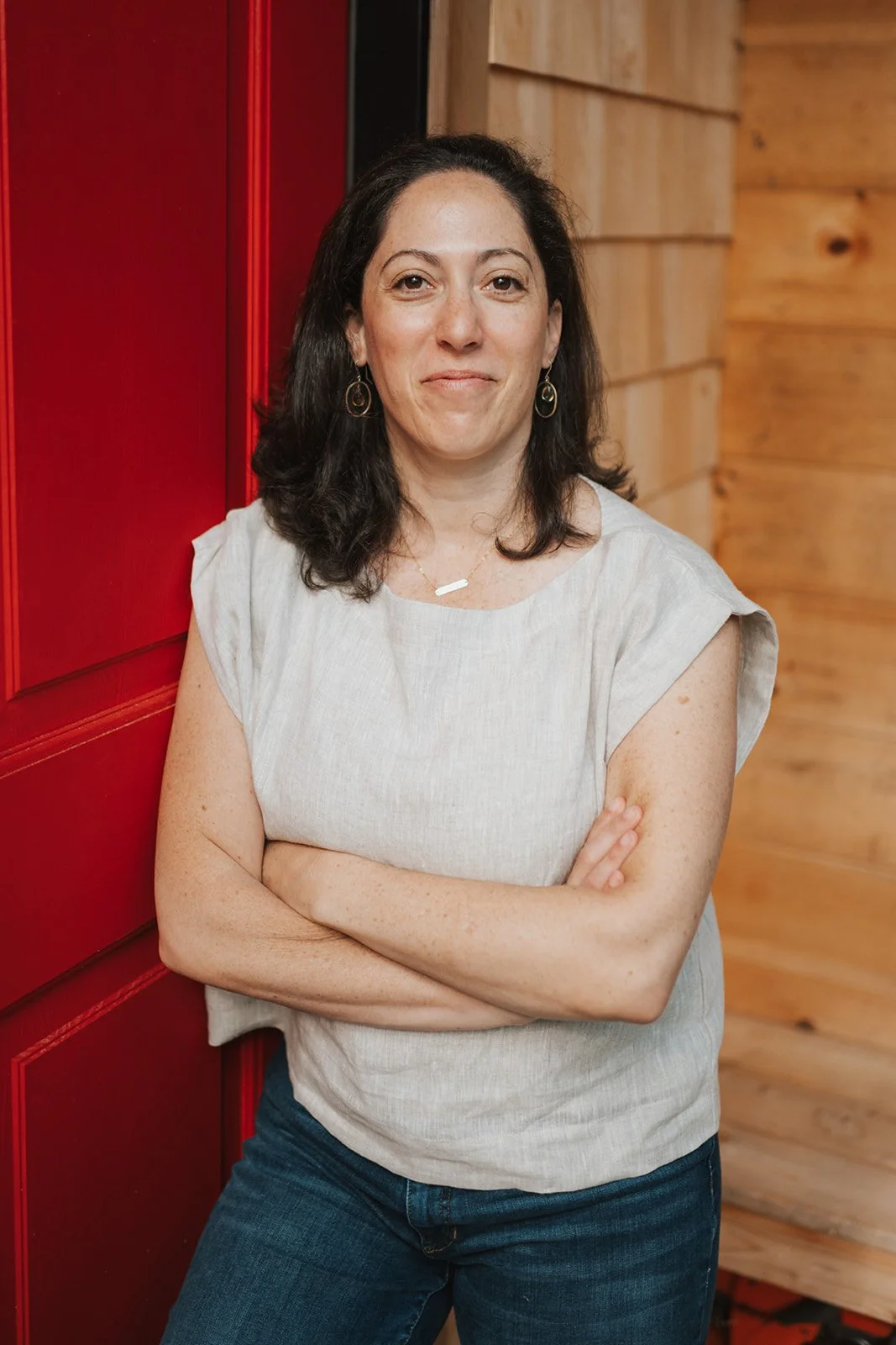 A woman with dark curly hair wearing a light beige sleeveless top and dark jeans, standing with arms crossed against a red door and wooden wall background.