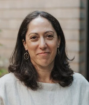 A woman with dark hair, wearing a light-colored top and hoop earrings, standing outdoors in front of a brick wall.