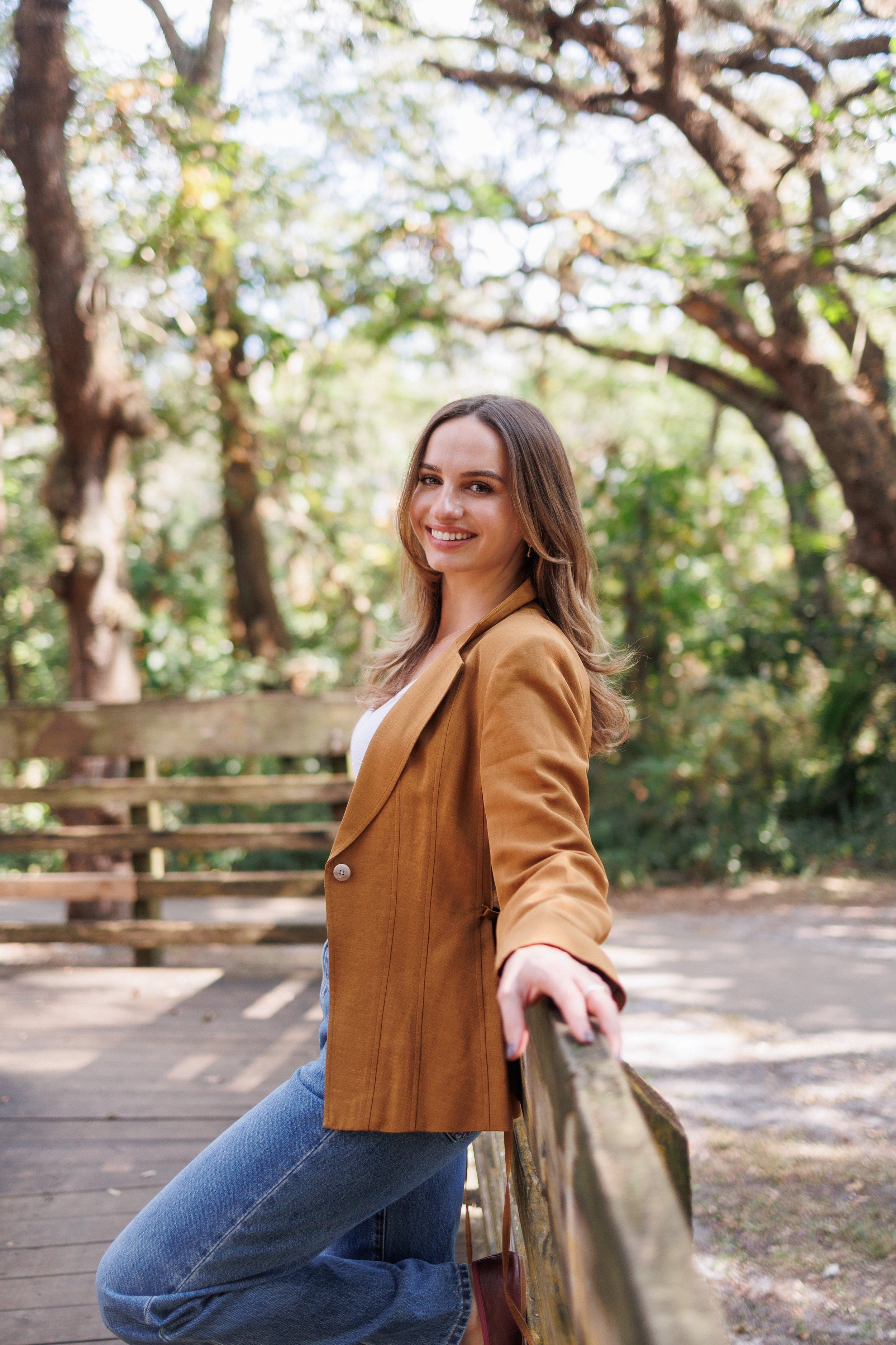 A female therapist with long brown hair and a bright smile, wearing a tan blazer over a white top and blue jeans, leaning on a wooden railing in a park surrounded by trees.