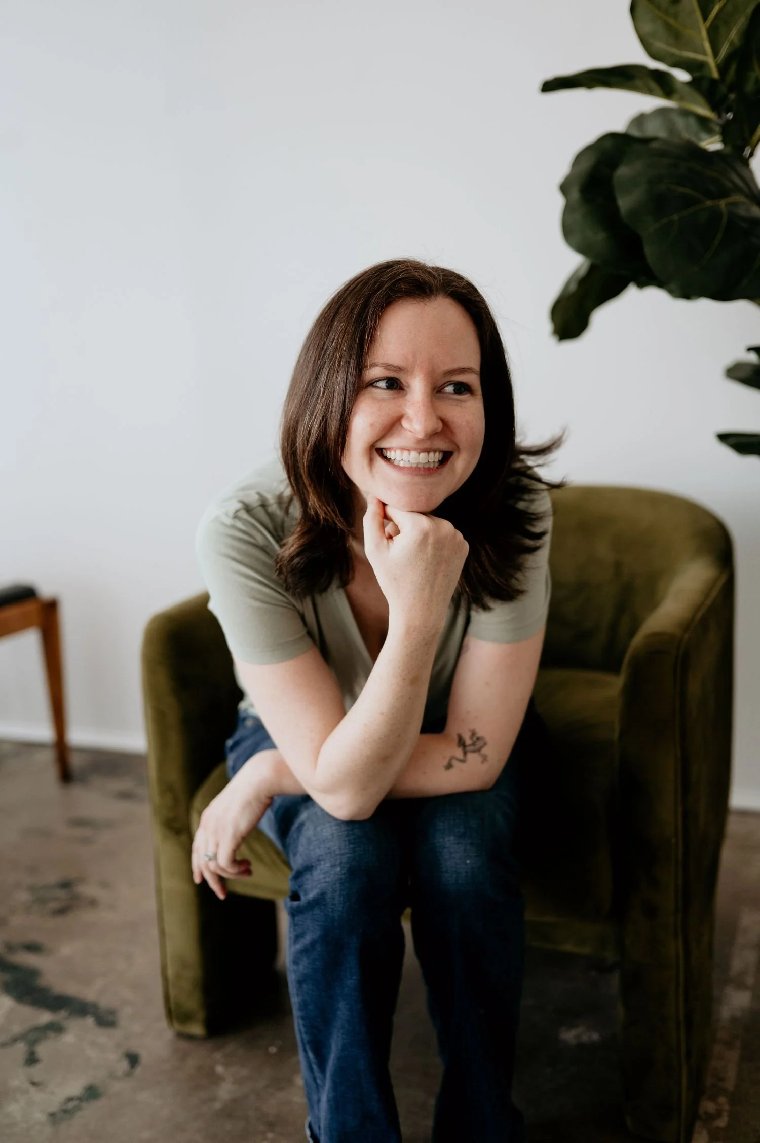 A woman sitting on an olive green armchair, smiling with her chin resting on her hand, indoors with a white wall and a large leafy plant nearby.