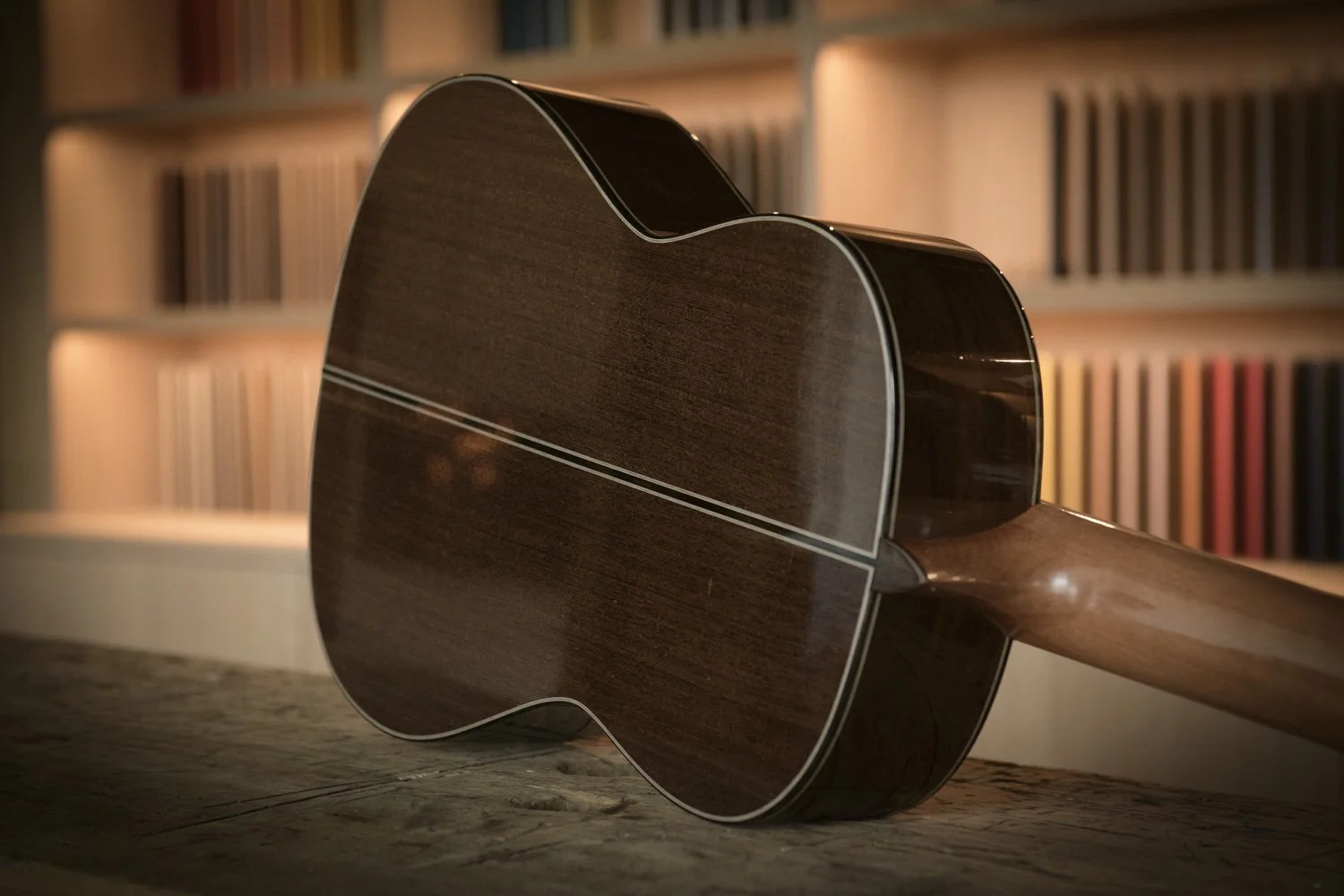 Back of a wooden acoustic guitar on a wooden surface with blurred bookshelf in the background.