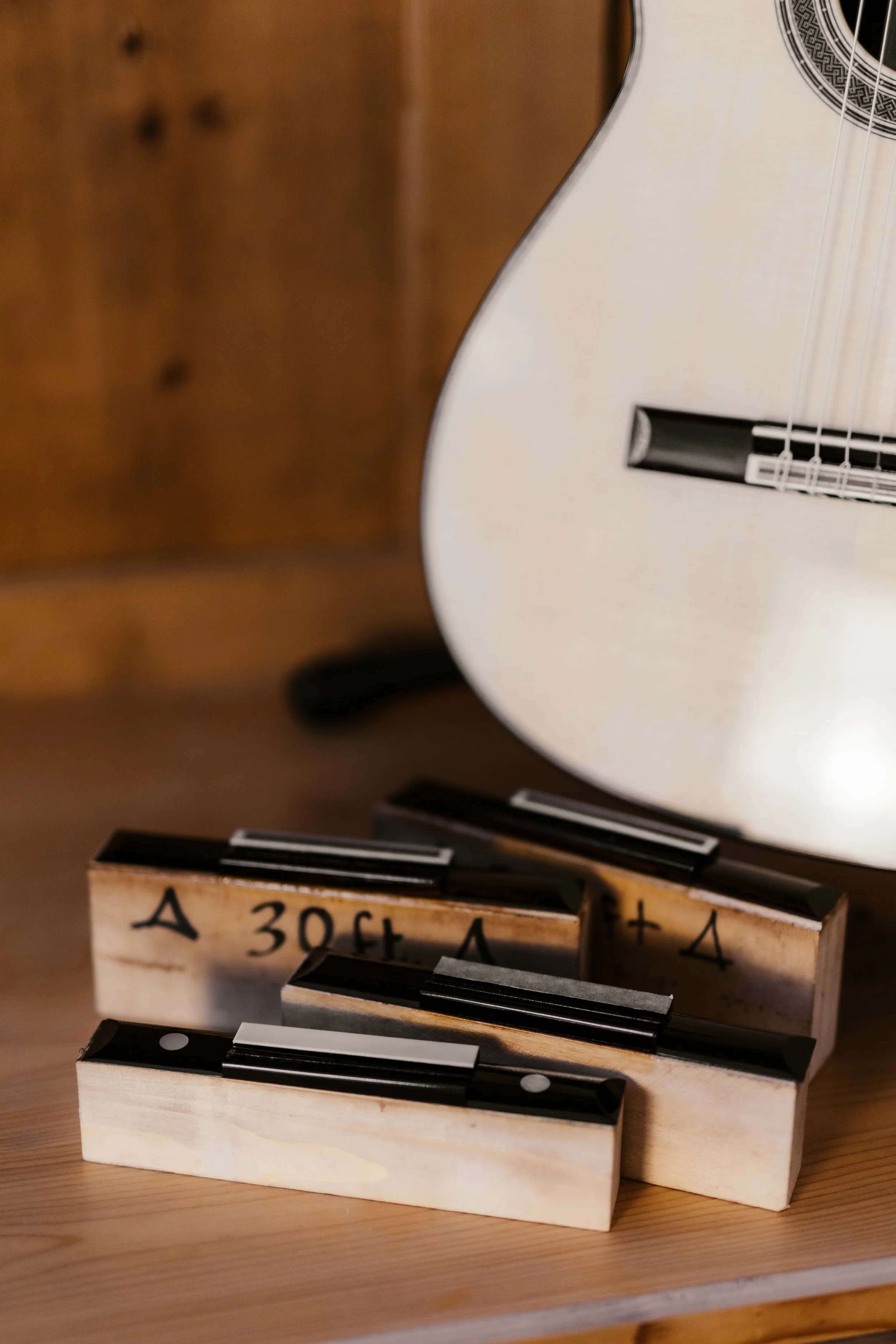 A white acoustic guitar partially visible behind a wooden surface with guitar slides or capos placed on it, some with handwritten markings.