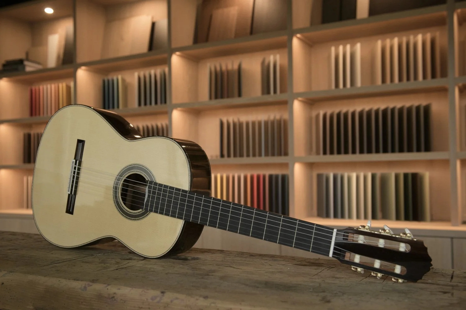 An acoustic guitar lying on a wooden table in front of a backlit bookshelf filled with colorful books.