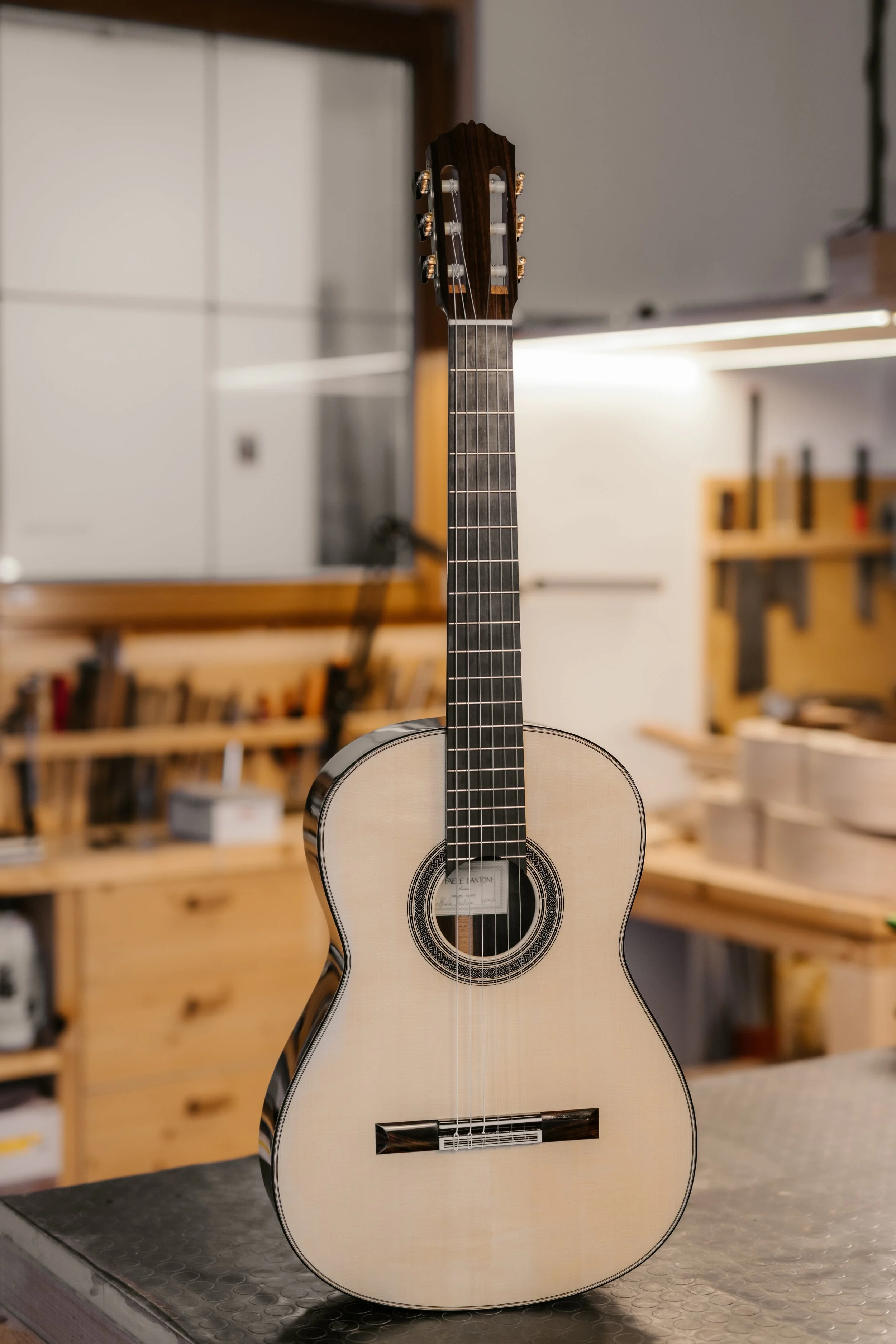 A classical guitar with a light-colored wooden body and a dark fretboard, standing upright in a workshop environment.