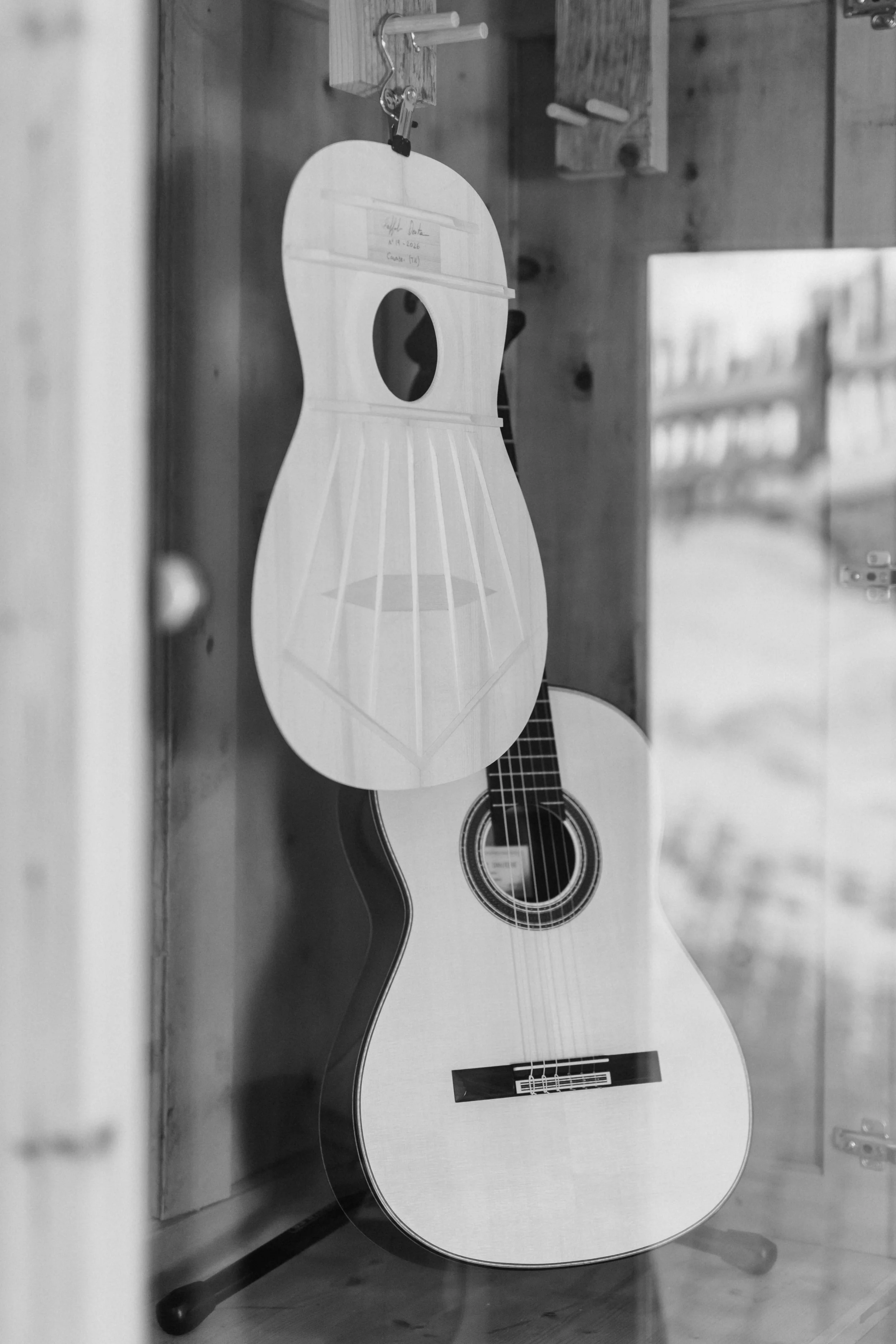 A classical guitar leaning against a wooden wall and a wooden binder clip holding a piece of paper with a wooden shape resembling a guitar body hanging above it.