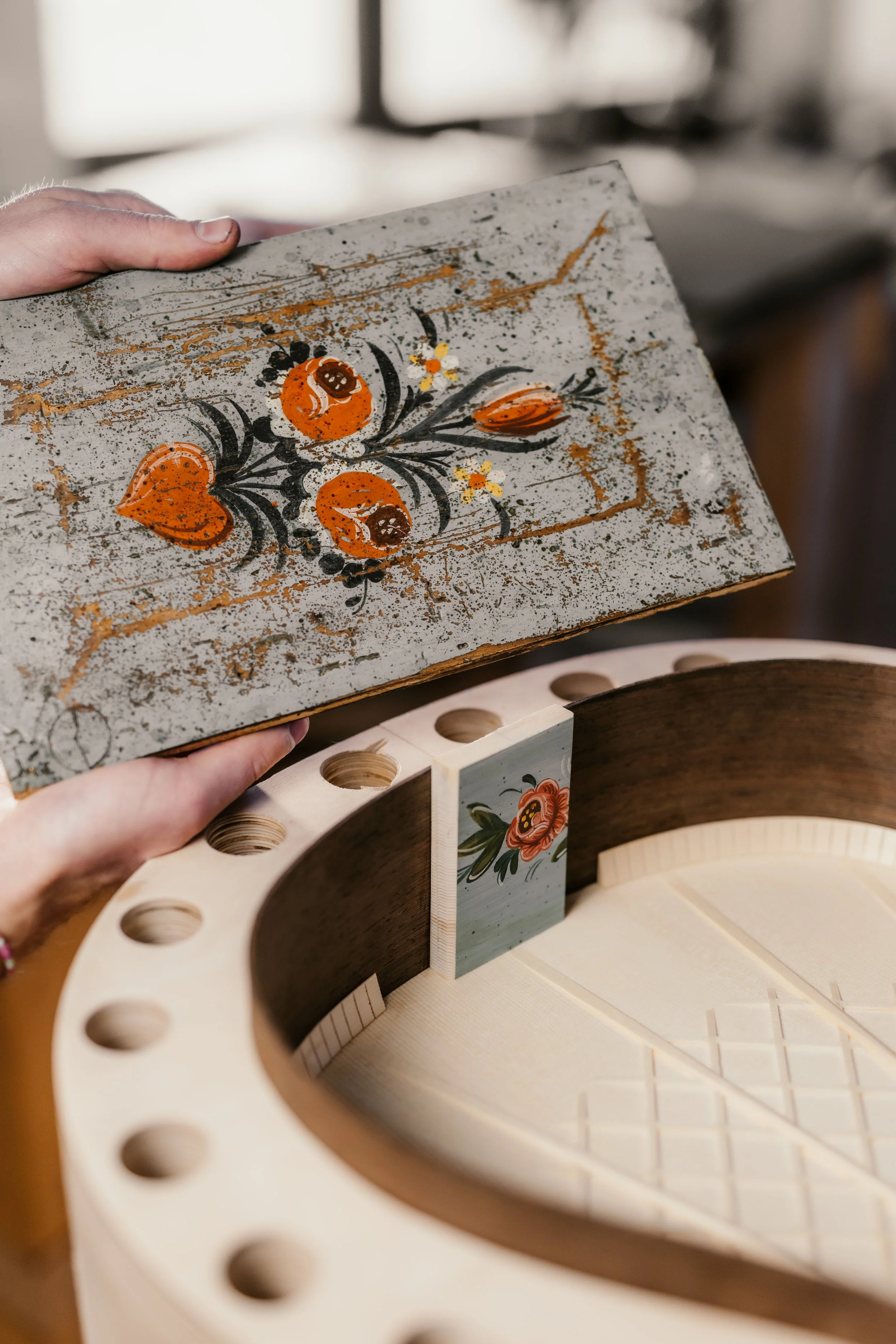 A person holding a wooden box with a painted floral design. The box is positioned above a circular wooden object with holes, possibly part of a musical instrument or craft project.
