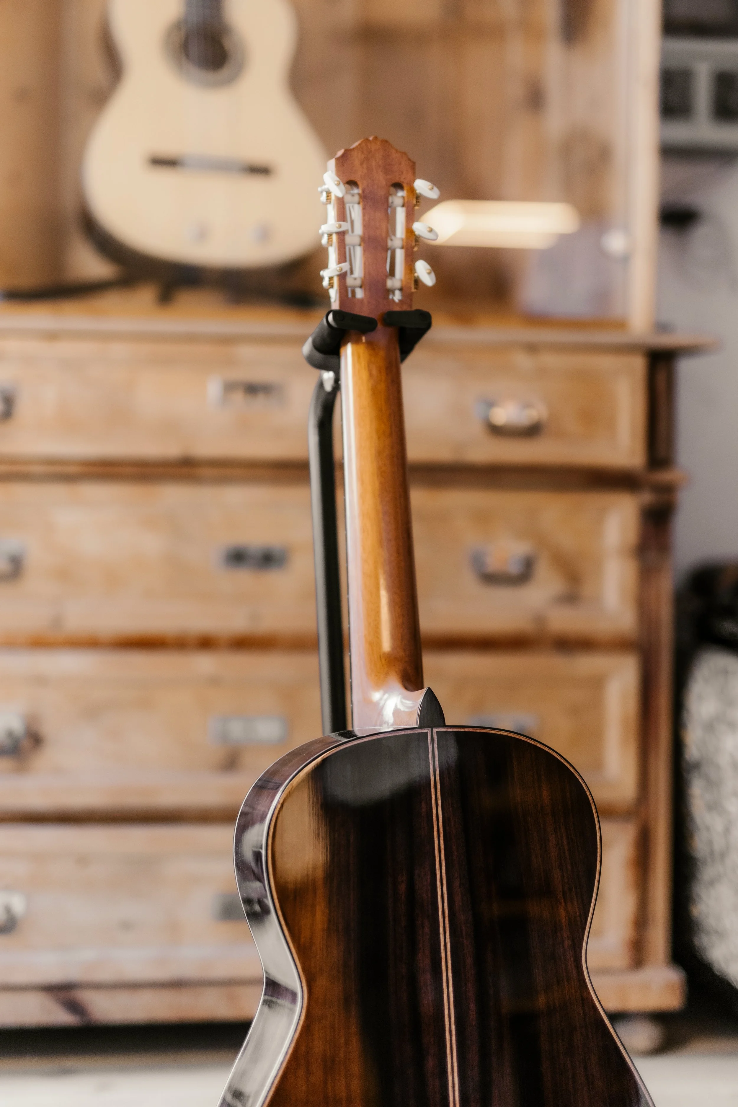 Back of a wooden acoustic guitar on a guitar stand in a room with wooden furniture and a second guitar hanging in the background.