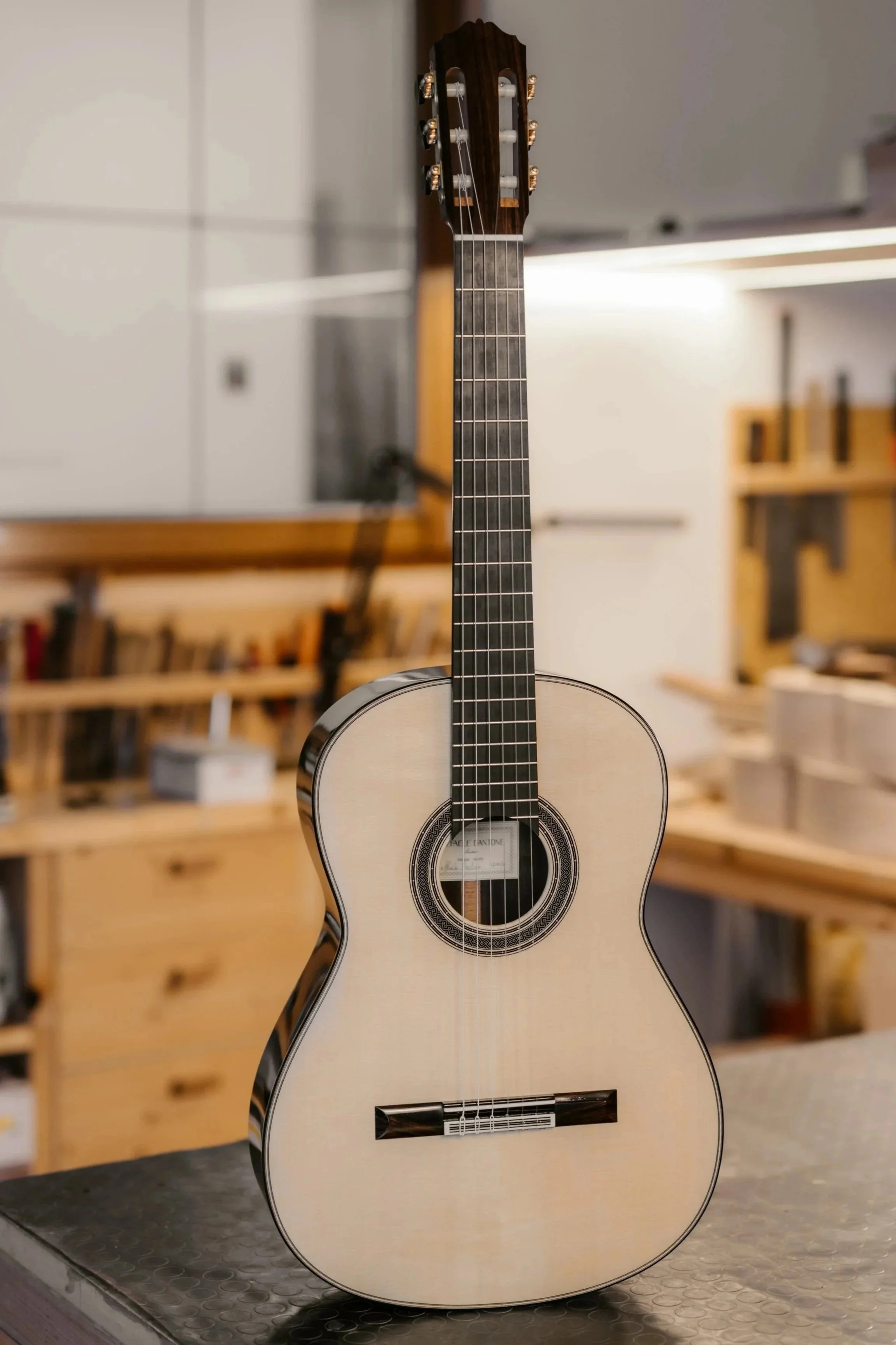 A light-colored acoustic guitar with a dark neck, standing upright on a table in a workshop or music studio.