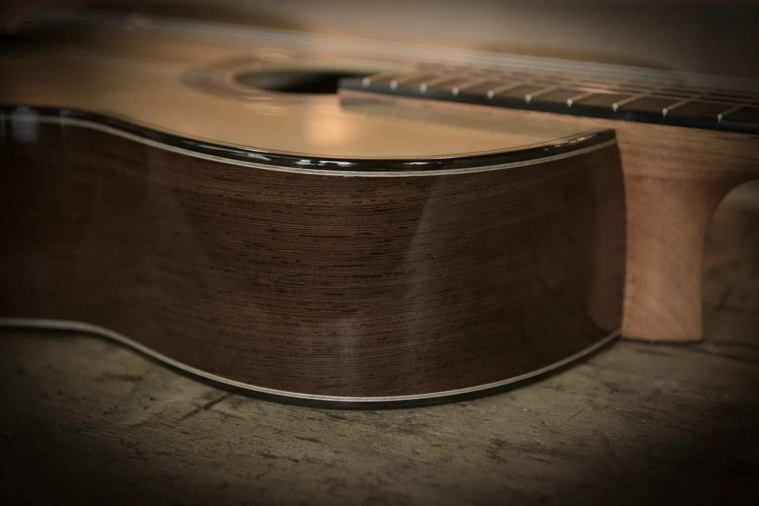 Close-up of an acoustic guitar resting on a wooden surface, showing the side and part of the top body with its wood grain detail.
