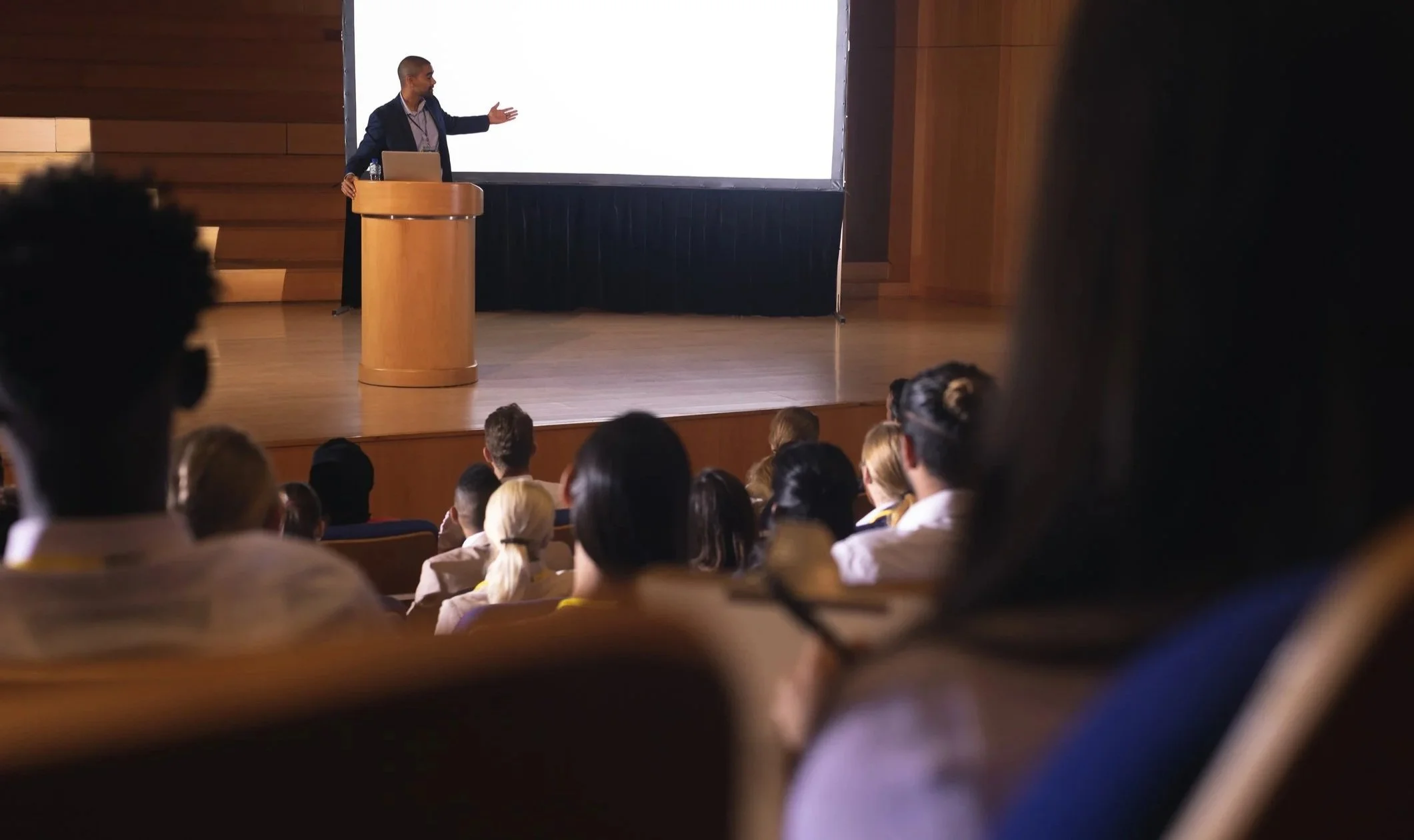 Man on stage pointing to white screen behind him as audience watches.