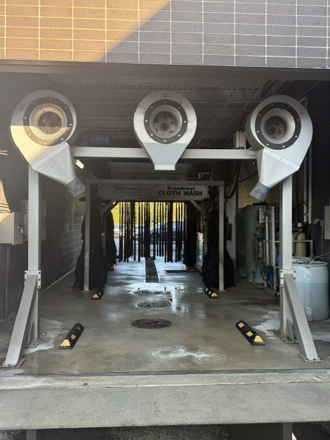 Automated car wash station with three large spray nozzles overhead and a pathway lined with black rollers for guiding vehicles.