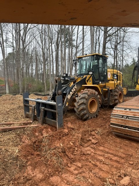A yellow SANY wheel loader with a black attachment, working on a construction site with dirt and trees in the background.