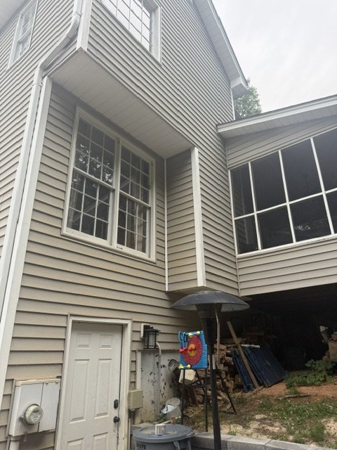 Back exterior of a house with beige vinyl siding, multiple large windows, and a small door. There is a patio umbrella, a colorful fish-shaped decoration, and some outdoor items underneath the house.