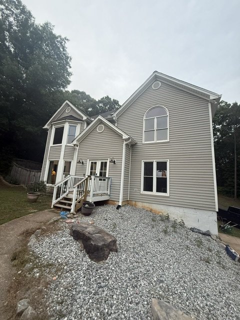 Back view of a beige two-story house with white trim, a deck with stairs, and gravel landscaping in the yard.