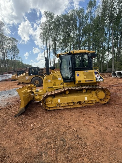 A yellow bulldozer at a construction site with other equipment and trees in the background.