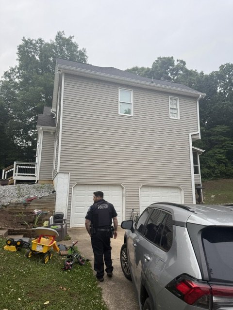 A police officer standing outside a gray house with a two-car garage, near a parked gray SUV, on a cloudy day. There are children's toys on the lawn.