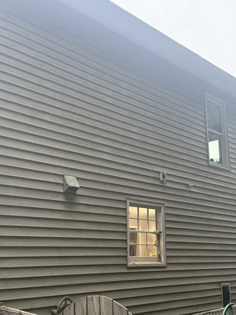 Close-up of a beige house exterior with horizontal vinyl siding, two small rectangular windows, and a small vent or fixture on the wall.