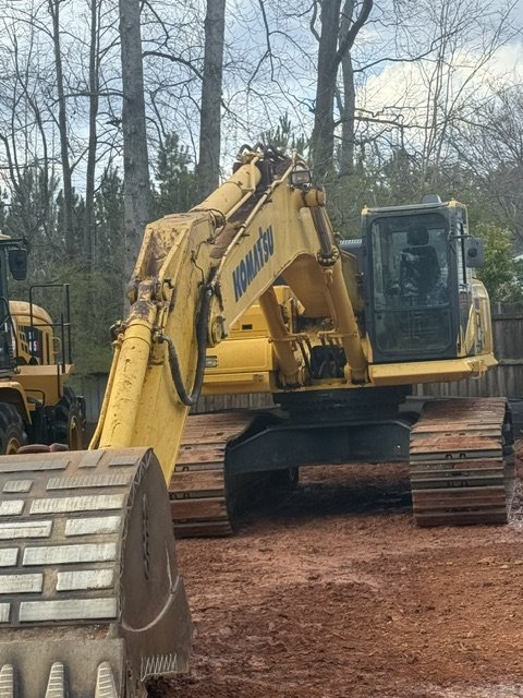 A yellow Komatsu excavator parked on a dirt lot with trees and a wooden fence in the background.