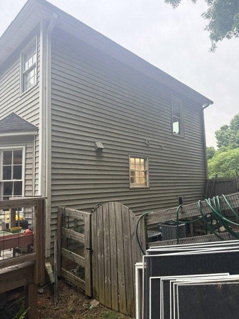 Back view of a gray two-story house with siding, showing two small windows and a light fixture. A wooden gate, outdoor furniture, and gardening tools are in the yard, along with trees and a cloudy sky.