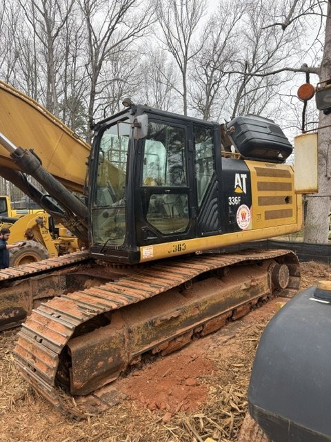 Yellow and black excavator with tracked wheels parked outdoors, trees and cloudy sky in the background.