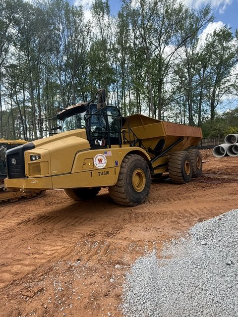 A yellow articulated dump truck at a construction site on dirt ground, with trees and pipes in the background.