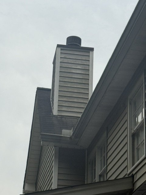 Side view of a house chimney and part of the roof with windows, under an overcast sky.