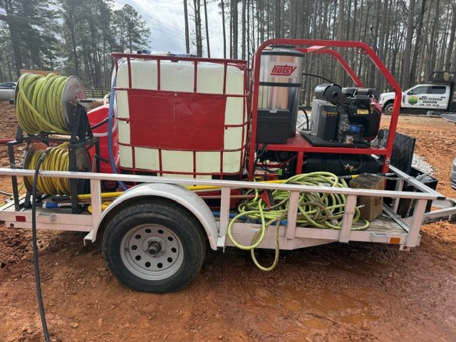 A mobile water or utility trailer equipped with hoses, a large water tank, and a pump system, parked on a dirt surface with trees in the background.
