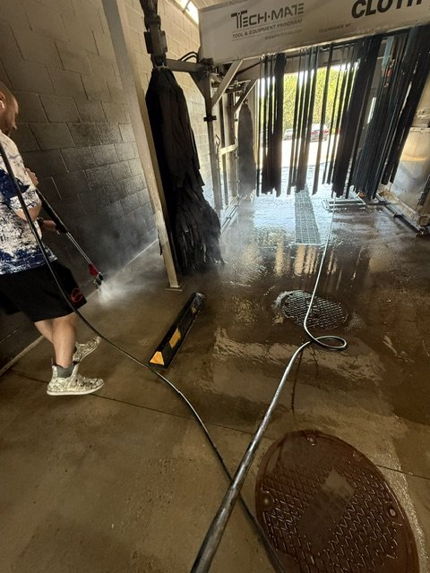 Person spraying water on the ground at an outdoor shoe cleaning station with hanging clothes in the background.