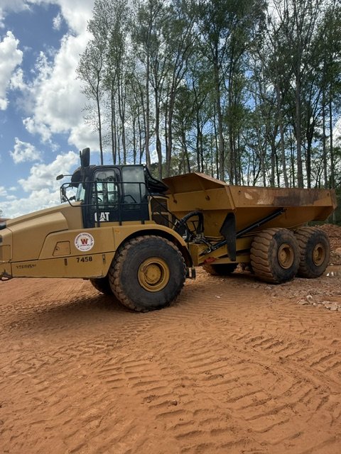A large yellow construction dump truck on dirt ground with trees and blue sky in the background.