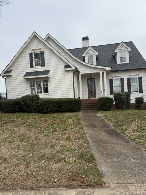 A two-story white house with black shutters, a gabled roof, and a brick front porch, surrounded by a grassy lawn and bushes.