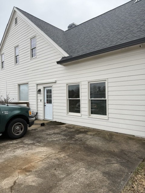 Exterior of a white house with a gable roof, three windows, a door with a window, and a black light fixture, with a green truck parked in front on a concrete driveway.