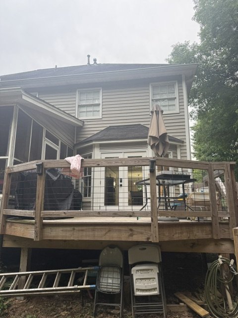 Backyard deck attached to a two-story house, with patio furniture and an umbrella, over storage area with a ladder and chairs underneath.