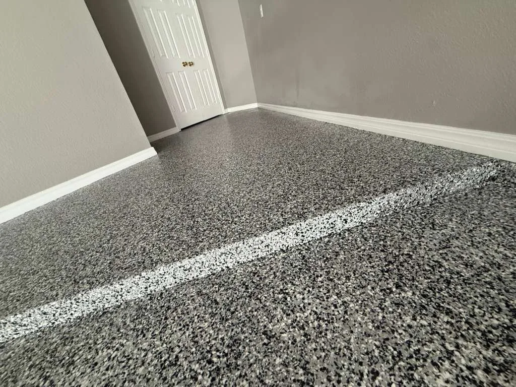 Close-up of a textured gray and black speckled epoxy floor with a white painted line, corner of a room with gray walls, white baseboards, and a closed white door with gold handles.