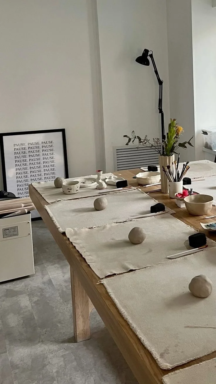 A dining table set for a meal, featuring ceramic bowls, black rectangular objects, a container with utensils, a vase with flowers, and place mats. A framed poster with repeated words 'PAUSE.' leans against the wall, and a black adjustable lamp is positioned behind the table.