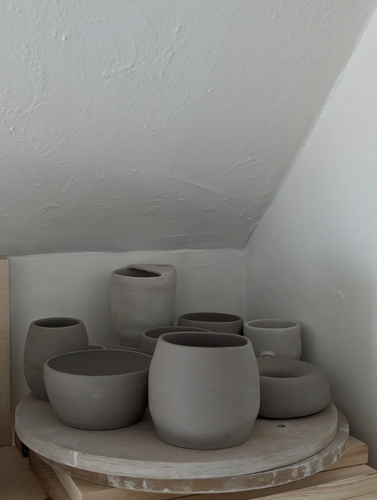 A collection of unglazed ceramic pots of varying shapes and sizes on a round wooden stand in a corner under a sloped ceiling.